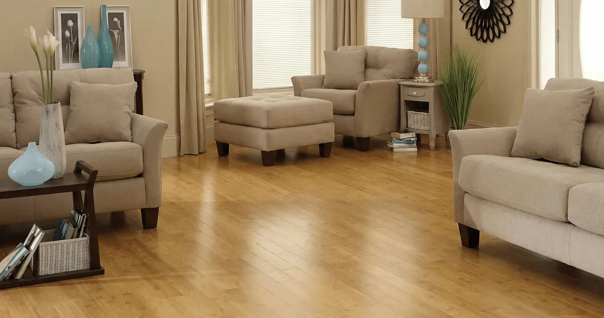 A living room with professionally installed bamboo flooring, beige sofas, a matching ottoman, a side table with a blue lamp, vases, framed pictures, and large windows with blinds and curtains in Chicago, IL